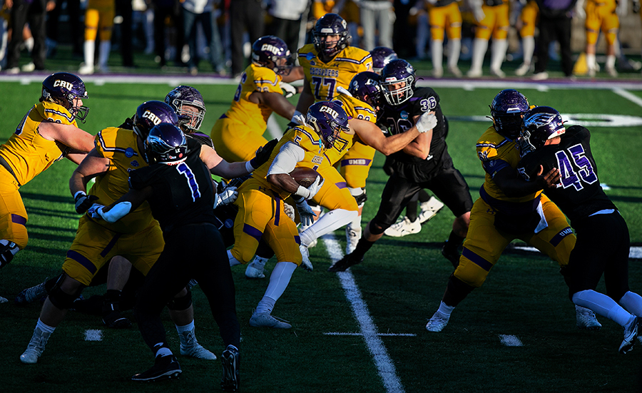 Warhawk Football in black uniforms plays defense against Mary Hardin-Baylor in yellow uniforms on the football field at Perkins Stadium.